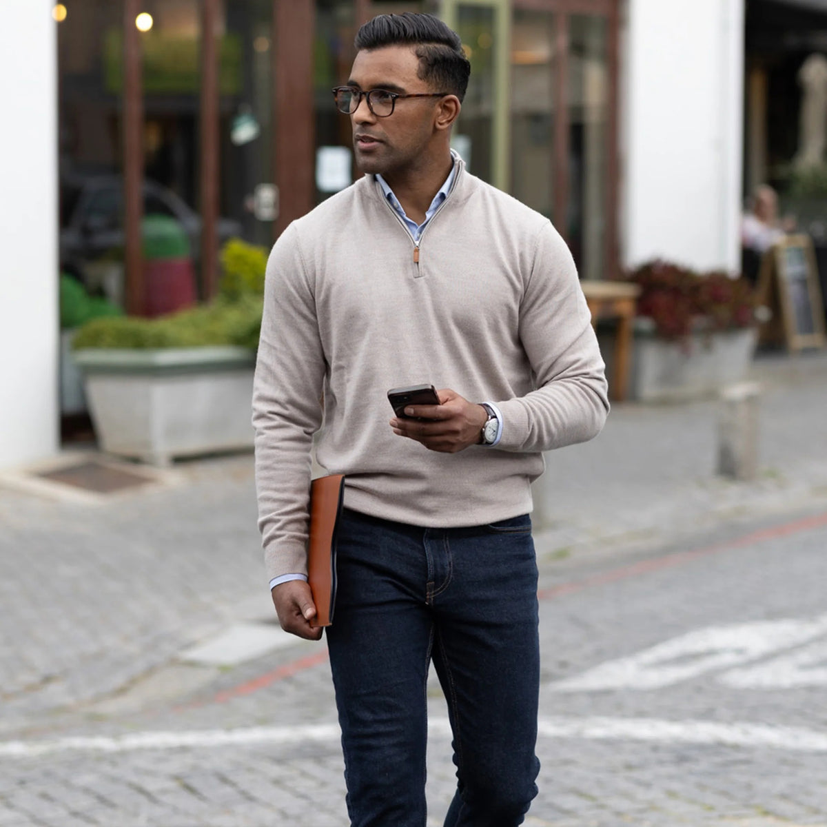 A man in glasses, wearing the COLLARS AND CO Cashmere Touch Australian Merino Wool 1/4 Zip Sweater in Oatmeal and dark jeans, walks on a cobblestone street holding a phone and a brown folder, with buildings and plants in the background.