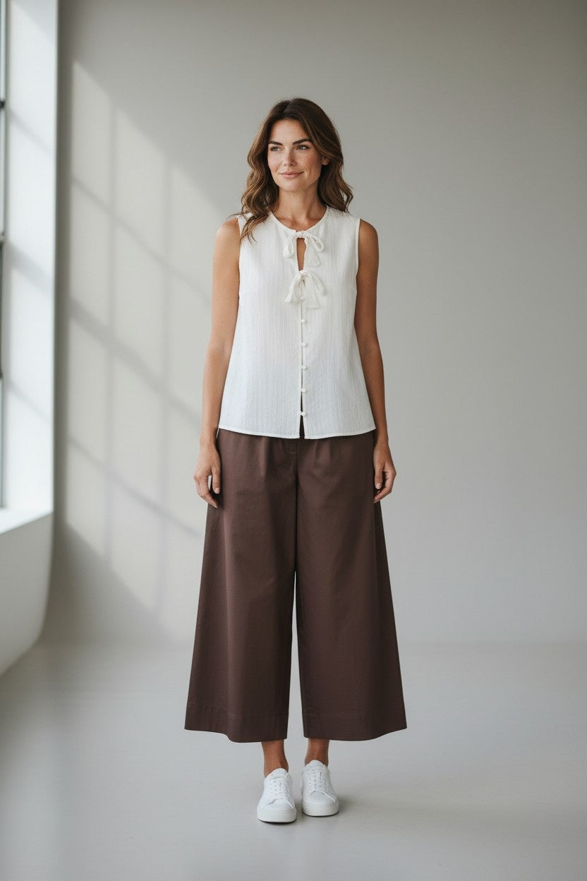 A woman stands indoors against a plain light gray background, wearing a SANDRO FERRONE Tie Front Sleeveless Top, wide-leg brown pants, and white sneakers.