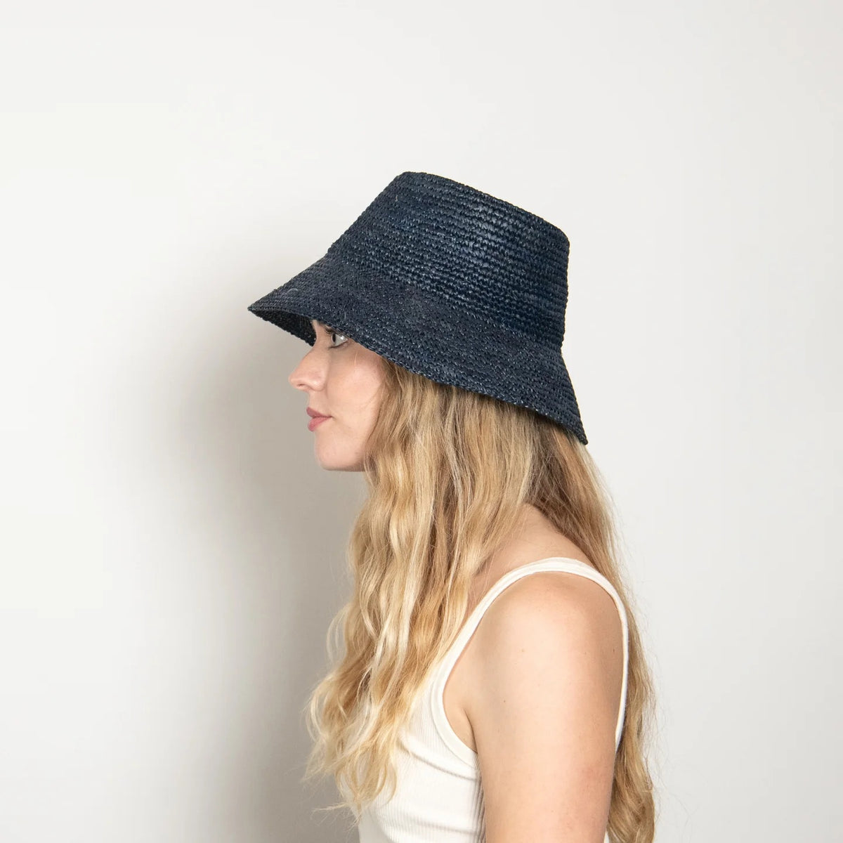 A woman with long, wavy blonde hair wears a sleeveless white top and the WYETH HATS - TALI Raffia Bucket Hat in Midnight by WYETH, featuring an adjustable fit, shown in profile against a plain light background.