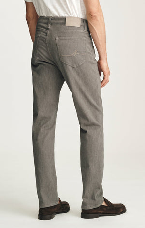 A man stands against a plain background wearing a dark brown sweater layered over a light collared shirt, paired with 34 HERITAGE - COURAGE STRAIGHT LEG PANTS IN STONE DIAGONAL by 34 HERITAGE, and brown shoes. One hand rests in his pocket and he maintains a neutral expression.