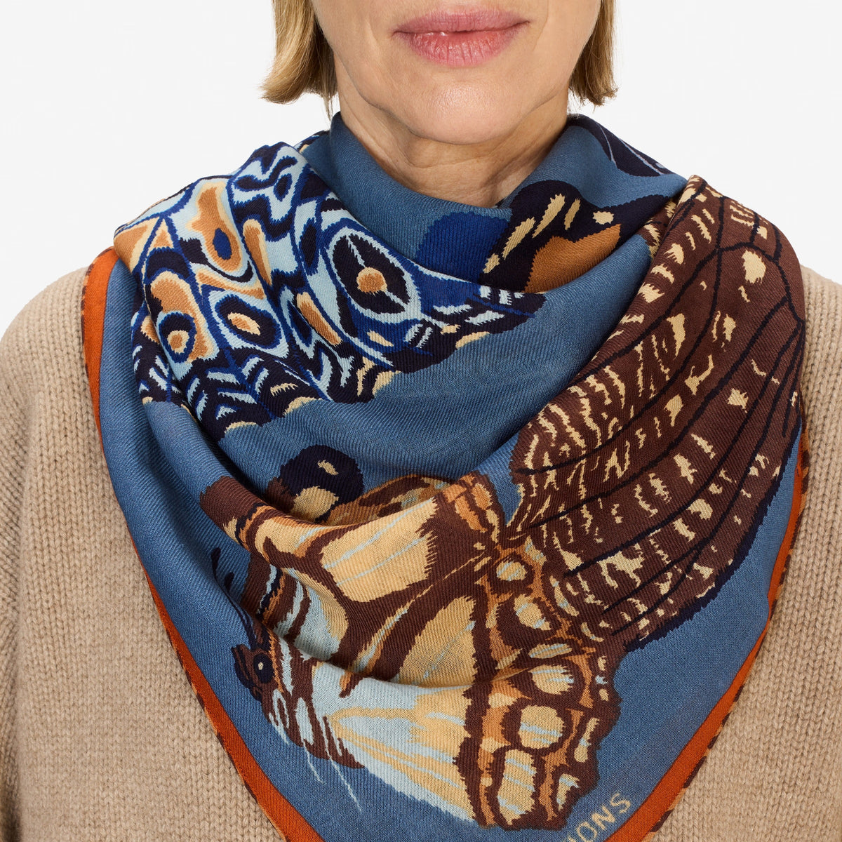 A woman with short blond hair and a beige sweater wears the INOUI EDITIONS PAPILLON (BUTTERFLIES) wool square scarf in duck blue, featuring colorful patterns, standing against a plain white background.