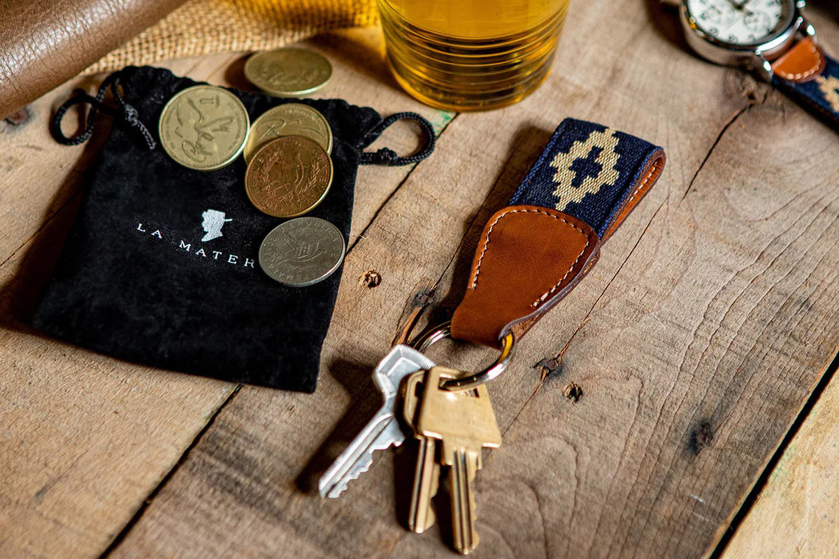 The LA MATERA - CORBINA WOVEN KEYCHAIN by LA MATERA features a silver metal ring, brown leather, and a navy blue strap with a beige diamond pattern inspired by Argentine artisan designs.