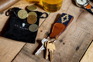 The LA MATERA - CORBINA WOVEN KEYCHAIN by LA MATERA features a silver metal ring, brown leather, and a navy blue strap with a beige diamond pattern inspired by Argentine artisan designs.