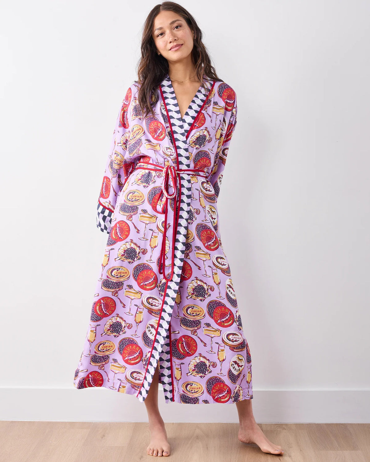 A woman stands barefoot against a white wall, smiling softly in the PRINTFRESH - ECO SATIN ROBE IN CHAMPAGNE AND CAVIAR by PRINTFRESH, featuring colorful food and champagne illustrations, checkered trim, and a matching waist tie.