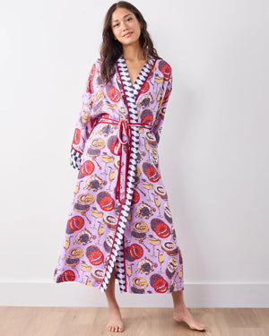 A woman stands barefoot against a white wall, smiling softly in the PRINTFRESH - ECO SATIN ROBE IN CHAMPAGNE AND CAVIAR by PRINTFRESH, featuring colorful food and champagne illustrations, checkered trim, and a matching waist tie.