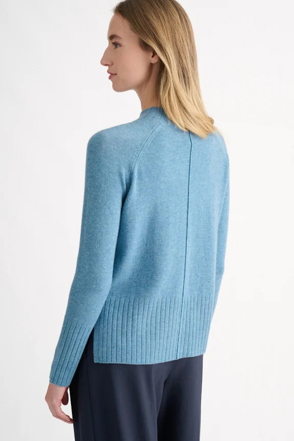 A woman with blonde hair models the KINROSS CASHMERE Hi Low Rib Trim Funnel in Loch—a light blue, long-sleeve cashmere pullover with ribbed trim—paired with dark pants. She stands against a plain white background, turned slightly to the side.