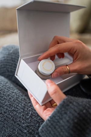 A person in a gray sweater holds an open white box containing MADISON GREEN’s “REST YOUR EYES ON THE COUCH,” lifting the small jar with a white and gold lid from the box—perhaps an essential oil blend designed for calming eye masks or aromatherapy eye covers.