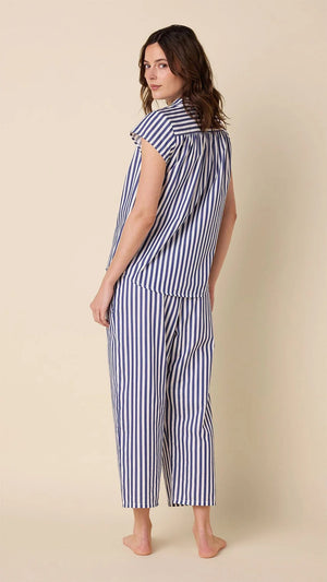 A woman wears CAT'S PAJAMAS - CLASSIC STRIPE LUXE PIMA CAPRI PAJAMAS with blue and white vertical stripes, short sleeves, and cropped pants, standing barefoot against a plain beige background.