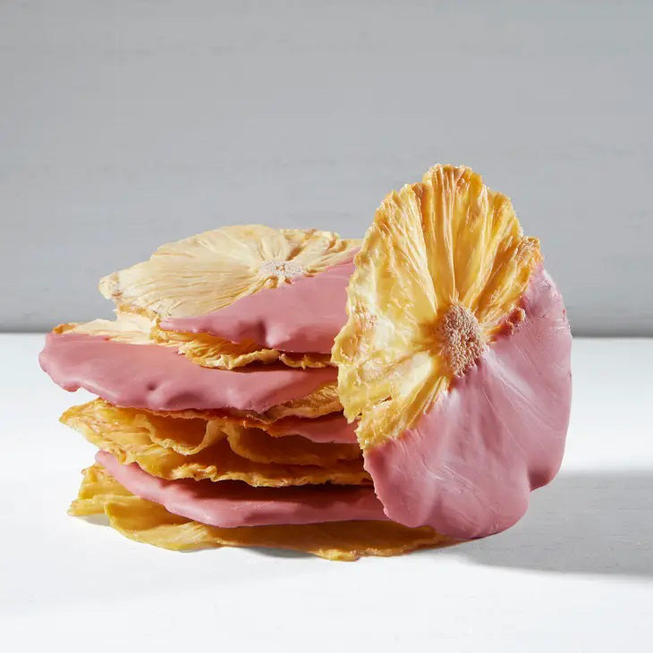 A clear plastic bag from DARDIMANS CALIFORNIA CRISPS contains several dried pineapple slices that are half-dipped in ruby chocolate. The label on the bag reads &quot;DARDIMANS - RUBY CHOCOLATE PINEAPPLE SLICES.&quot; The bag, brimming with tropical sweetness, is secured with a twist tie and set against a textured, light grey background.
