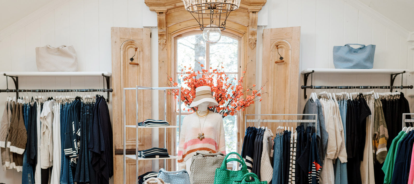A boutique clothing store displays neatly arranged racks of clothing, woven bags, and a mannequin wearing a hat and sweater in front of a rustic window with orange flowers and natural light streaming in.