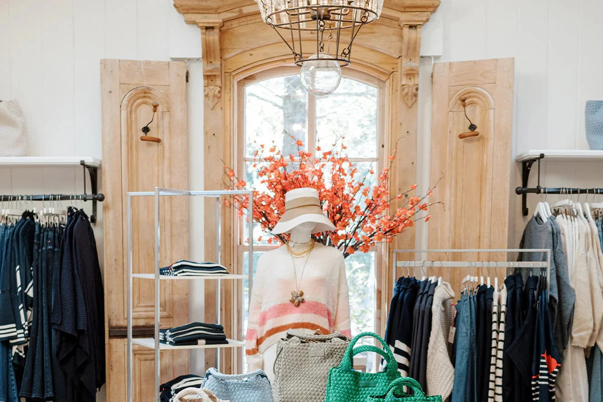 A boutique clothing store display features a mannequin in a sweater and hat, surrounded by folded clothes, green bags, and a floral arrangement, with wooden doors and a large window in the background.