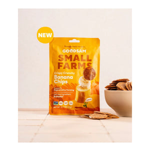 A yellow bag of GOODSAM FOODS GOODSAM - BANANA CHIPS stands upright on a light surface beside a bowl of banana chips, with a “NEW” label in the top left highlighting women-led innovation for healthy snacks.