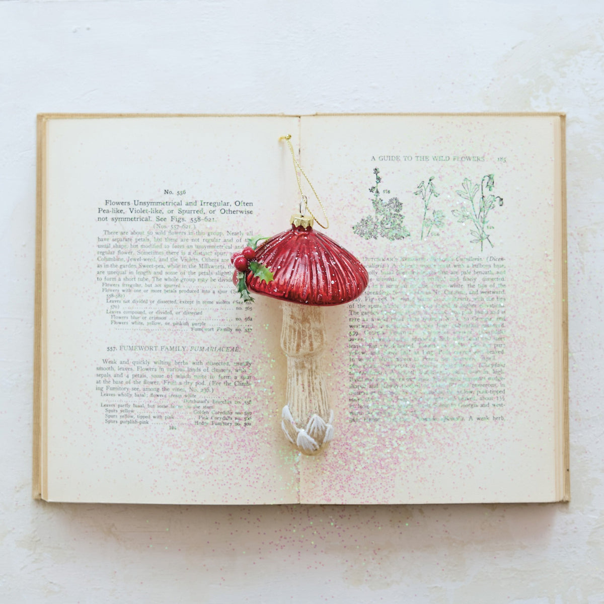 A MUSHROOM ORNAMENT WITH HOLLY AND GLITTER shaped like a glass mushroom with a shiny red cap, a textured off-white stem, and a small sprig of holly on top.