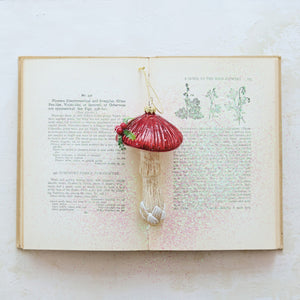 A MUSHROOM ORNAMENT WITH HOLLY AND GLITTER shaped like a glass mushroom with a shiny red cap, a textured off-white stem, and a small sprig of holly on top.