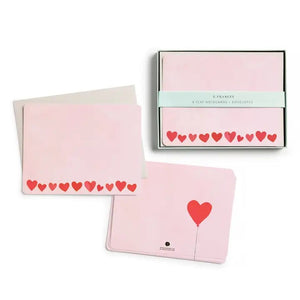 The E. FRANCES - FLAT NOTES BLUSH MINI HEARTS by E. FRANCES PAPER are pale pink notecards with rounded corners and red and pink hearts along the bottom, stacked atop light gray envelopes.