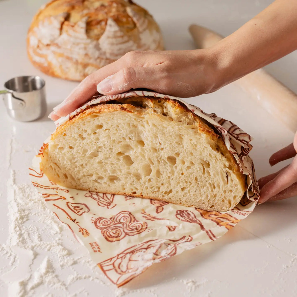 A loaf of bread is partially tucked inside the BEES WRAP- BEESWAX REUSABLE BREAD BAKERY LANE bag by BEES WRAP, featuring a small white label and displayed against a plain white background—a perfect choice for a plastic-free kitchen.