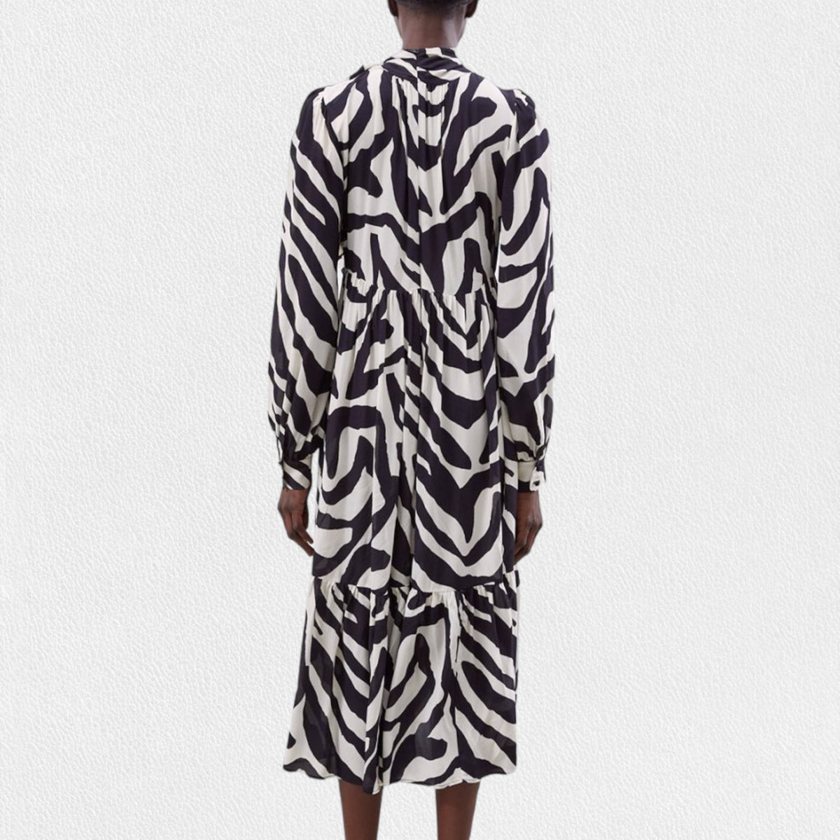 A person is wearing the MEIMEIJ Ruffle Print Dress, a long-sleeved, black and white zebra print midi dress made from 100% viscose fabric. It features a high neckline, gathered waist, tiered skirt, and buttoned cuffs. The background is plain white.
