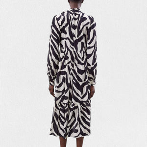 A person is wearing the MEIMEIJ Ruffle Print Dress, a long-sleeved, black and white zebra print midi dress made from 100% viscose fabric. It features a high neckline, gathered waist, tiered skirt, and buttoned cuffs. The background is plain white.