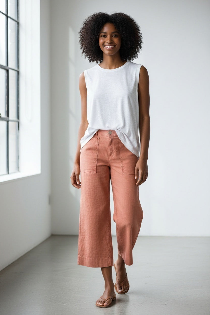 A person wearing the FRANK &amp; EILEEN Limerick Cropped Wide Leg Carpenter Pant in Italian Performance Linen Canyon, paired with a white top and tan sandals, stands on outdoor stone steps with hands in pockets; the background features grass, rocks, and sunlight.