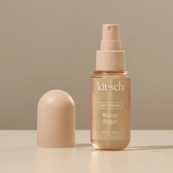 A bottle of KITSCH - HAIR PERFUME MIST IN WARM SUGAR stands between a slice of cake and blue cotton candy, with labeled fragrance notes: pink grapefruit, whipped cream, spun sugar, bergamot blossom, musk, vanilla, and praline. This odor-eliminating formula from KITSCH ensures your hair smells irresistibly sweet and fresh all day.