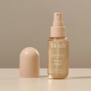 A bottle of KITSCH - HAIR PERFUME MIST IN WARM SUGAR stands between a slice of cake and blue cotton candy, with labeled fragrance notes: pink grapefruit, whipped cream, spun sugar, bergamot blossom, musk, vanilla, and praline. This odor-eliminating formula from KITSCH ensures your hair smells irresistibly sweet and fresh all day.