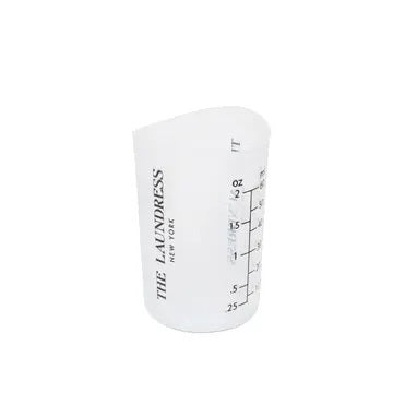 A hand pours liquid from THE LAUNDRESS - MEASURING CUP, a compact white silicone vessel clearly marked up to 2 ounces. The text highlights its use for detergent, machine washability, and a flexible yet sturdy design—ideal for precise fabric care by THE LAUNDRESS.