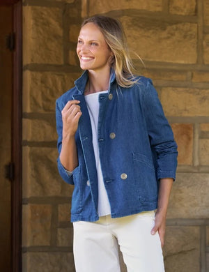 A woman stands smiling in front of a stone wall, wearing the FRANK & EILEEN Mini Belfast Cropped English Peacoat in 1984 Wash Denim over a white top and white pants.