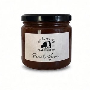 The FEARRINGTON SIGNATURE COLLECTION - FEARRINGTON HOUSEMADE PEACH JAM comes in a glass jar featuring a black lid and is adorned with a black and white striped ribbon. The white label reads "Peach Jam" and lists the ingredients as peach, vanilla, lemon, pectin, and sugar. Perfect as a hostess gift, it also notes that it is handmade by FEARRINGTON HOUSE and may contain allergens.