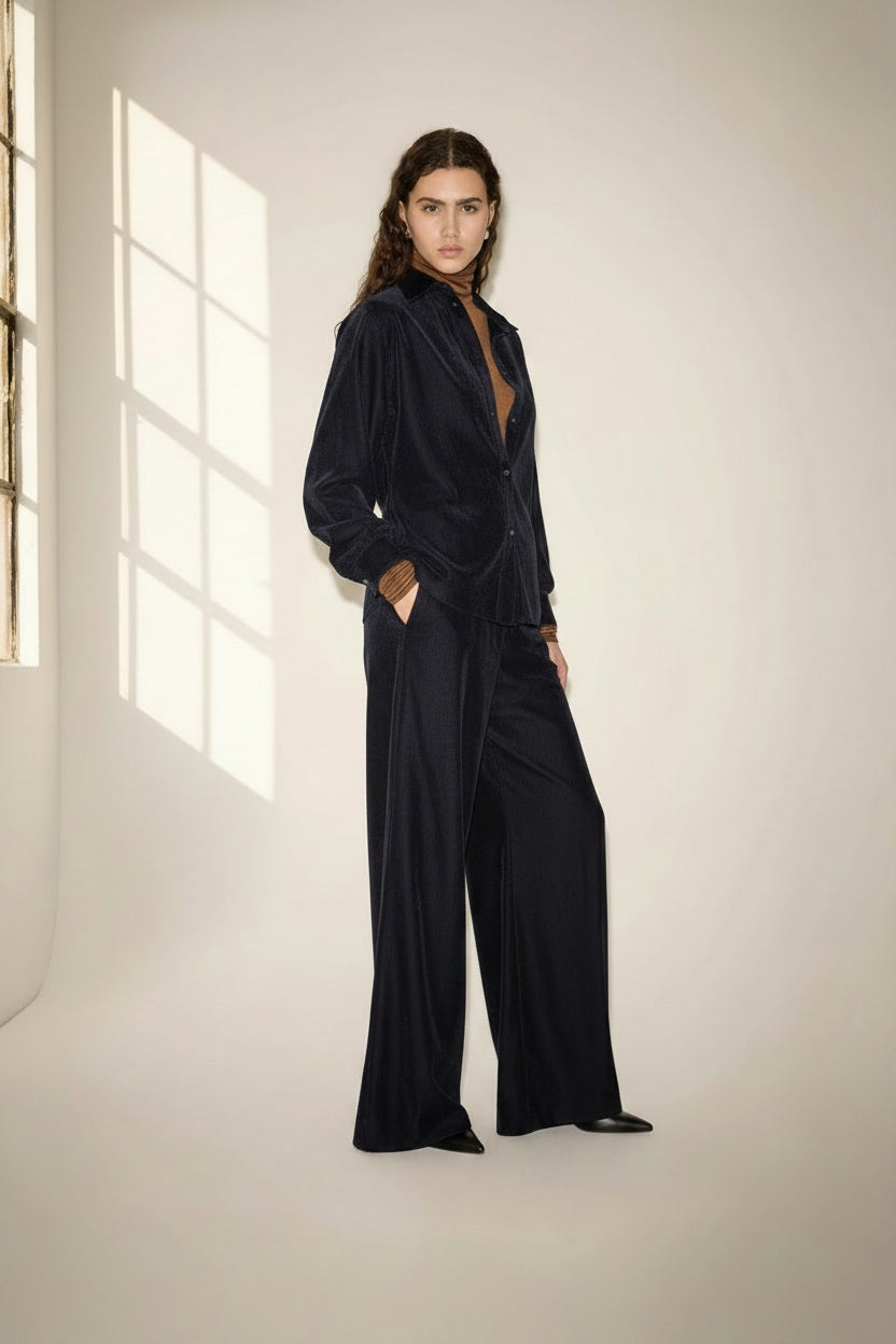 A person with long, wavy brown hair wears a loose-fitting, dark velvet suit over a brown top and stands in a softly-lit room with a large window casting light and shadows on the wall. The look is completed with NUDE’s Wide Leg Trouser in Blue, adding an effortless elegance to the ensemble.