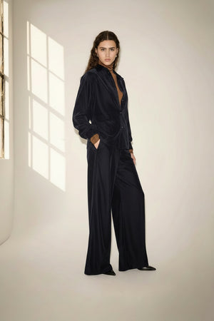 A person with long, wavy brown hair wears a loose-fitting, dark velvet suit over a brown top and stands in a softly-lit room with a large window casting light and shadows on the wall. The look is completed with NUDE’s Wide Leg Trouser in Blue, adding an effortless elegance to the ensemble.