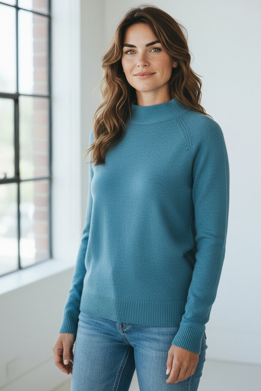 A woman with long brown hair wearing the KINROSS Cashmere Garter Funnel Cotton Sweater in Loch stands indoors near a large window, smiling softly at the camera.