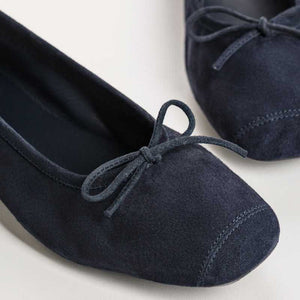 Someone stands on a light floor, one foot slightly raised, wearing REQINS - HARMONY suede ballerina flats in marine with a small bow, paired with blue jeans featuring frayed hems.