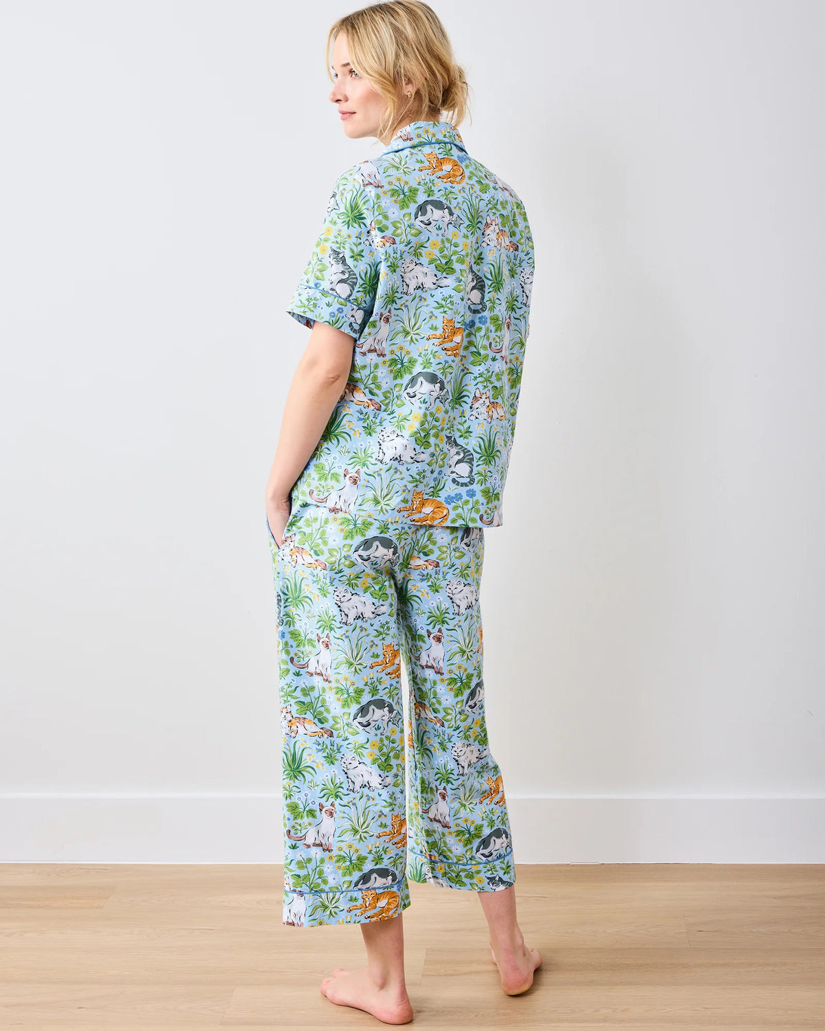 Wearing PRINTFRESH&#39;s &quot;Must Love Cats&quot; cropped pajama set, made of organic cotton poplin, a person stands barefoot against a white wall. The colorful, cat-themed design features short sleeves and cropped pants as they smile slightly.