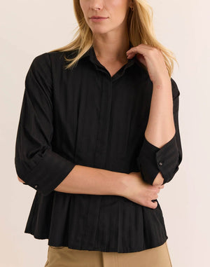 A person wearing the HINSON WU - MARTINA 3/4 SLEEVE SHIRT IN CLASSIC STRIPE BLACK, a button-down blouse with subtle vertical stripes by HINSON WU, paired with beige pants, stands against a plain light background.