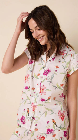 A person stands barefoot, wearing the CAT'S PAJAMAS - ROSA AND BIRD LUXE PIMA CAPRI PAJAMA SET from CAT'S PAJAMAS, featuring pink, purple, and green florals on a cream 100% cotton base with short sleeves and cropped pants. The backdrop is light beige.