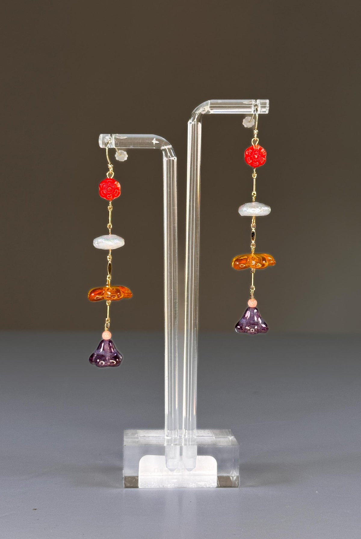 SANDRINE FRANCE STUDIO - FLEUR EARRINGS are shown on a clear stand, with red beads, white discs, orange beads, and purple glass flowers. Each piece is handmade in NYC and set against a plain gray background.