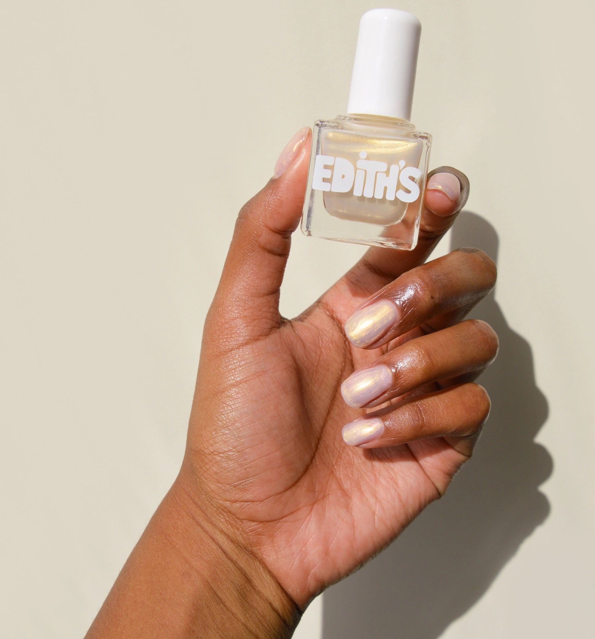 A hand with shimmering pearl nail polish holds a clear bottle of TENOVERTEN - LUXE AND BAGELS NAIL POLISH by TEN OVER TEN against a pale background.