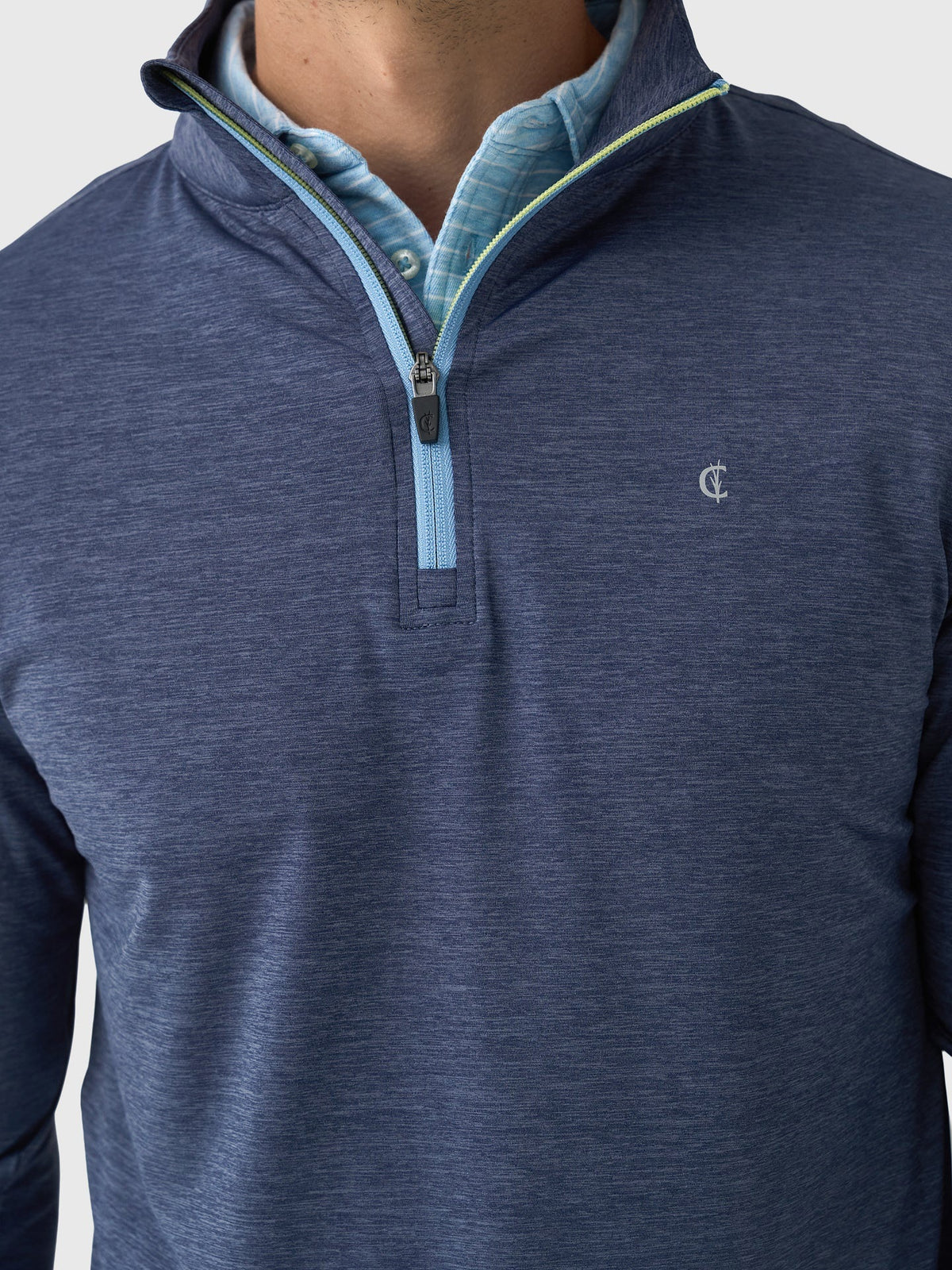 A person wears a CRIQUET - FEATHER PERFORMANCE KNIT QUARTER ZIP PULLOVER over a light blue checkered shirt. The moisture-wicking brrr° pullover by CRIQUET features a small embroidered logo on the left chest. Only the upper torso and neck are visible.