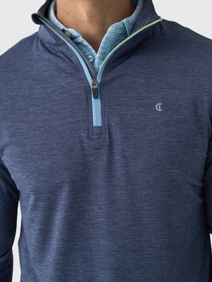 A person wears a CRIQUET - FEATHER PERFORMANCE KNIT QUARTER ZIP PULLOVER over a light blue checkered shirt. The moisture-wicking brrr° pullover by CRIQUET features a small embroidered logo on the left chest. Only the upper torso and neck are visible.