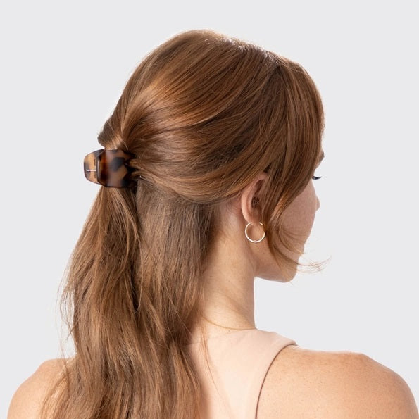 A woman with light skin and reddish-brown hair styled in a low bun, secured with the KITSCH - ECO FRIENDLY MEDIUM CLAW CLIP (2 PIECE SET), wears a beige top and touches her neck with manicured nails, shown in profile against a plain light background.