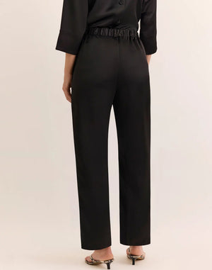 A person is wearing HINSON WU’s HOSTESS LIQUID SATIN PANT IN BLACK—high-waisted, wide-leg pants crafted from lustrous liquid satin fabric—paired with a black three-quarter sleeve top and open-toe heels with a thin strap. Their left hand is in the pocket of the pants, and their head is not visible.