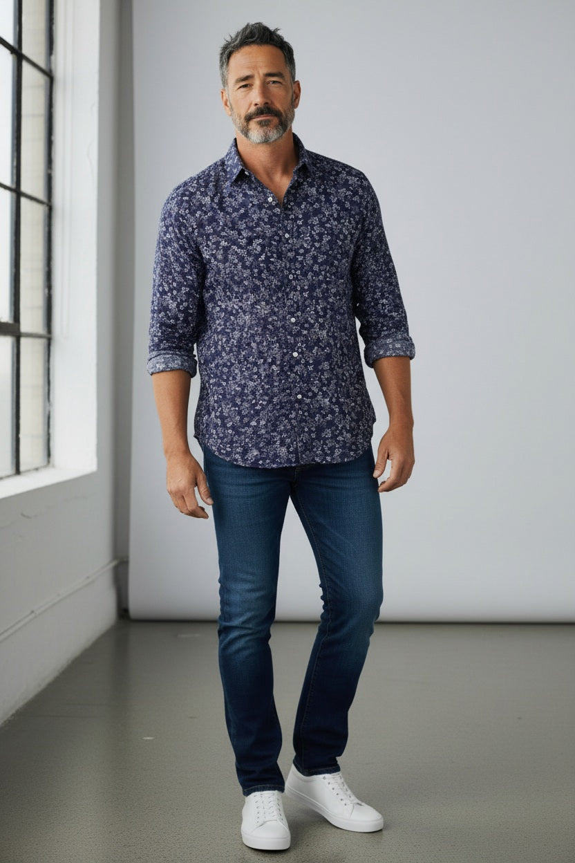 A man with gray hair and a beard wears the FRANK &amp; EILEEN – FINBAR Men’s Button Up Shirt in Italian Chambray, paired with dark blue jeans and white sneakers, standing indoors by a large window and light gray wall.
