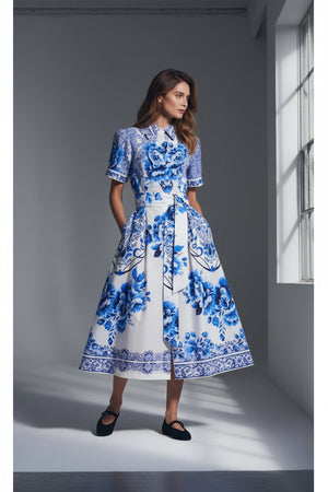 A woman with straight, shoulder-length hair wears the MILVA MI - COTTON FLORA PRINT DRESS IN BLUE AND WHITE by MILVA, featuring blue and white floral patterns. She faces away in flat black shoes against a plain gray background.