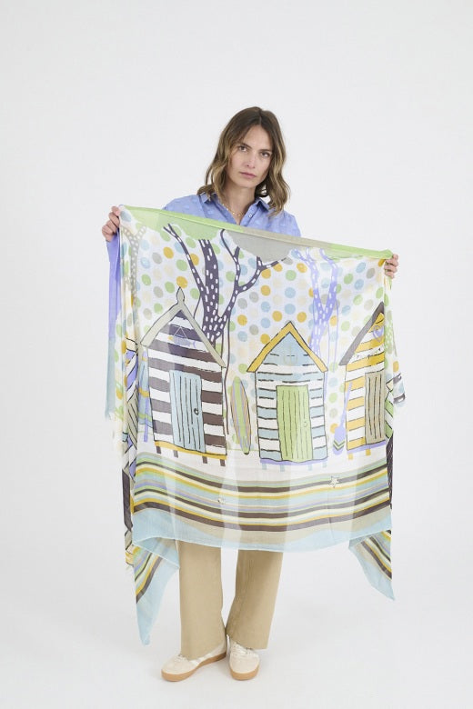 A woman in a light blue textured blouse and beige pants stands with one hand in her pocket, wearing the colorful STORIATIPIC - HEIDI SCARF from STORIATIPIC, its chic cotton fabric billowing stylishly around her neck.