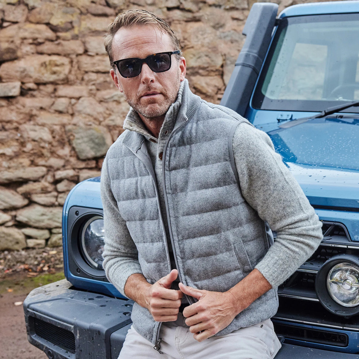 A smiling man in a light blue shirt and the COLLARS AND CO - REGENT REVERSIBLE QUILTED WOOL VEST holds a blue umbrella on a rainy street—showcasing COLLARS AND CO's ideal insulated layering for wet days.