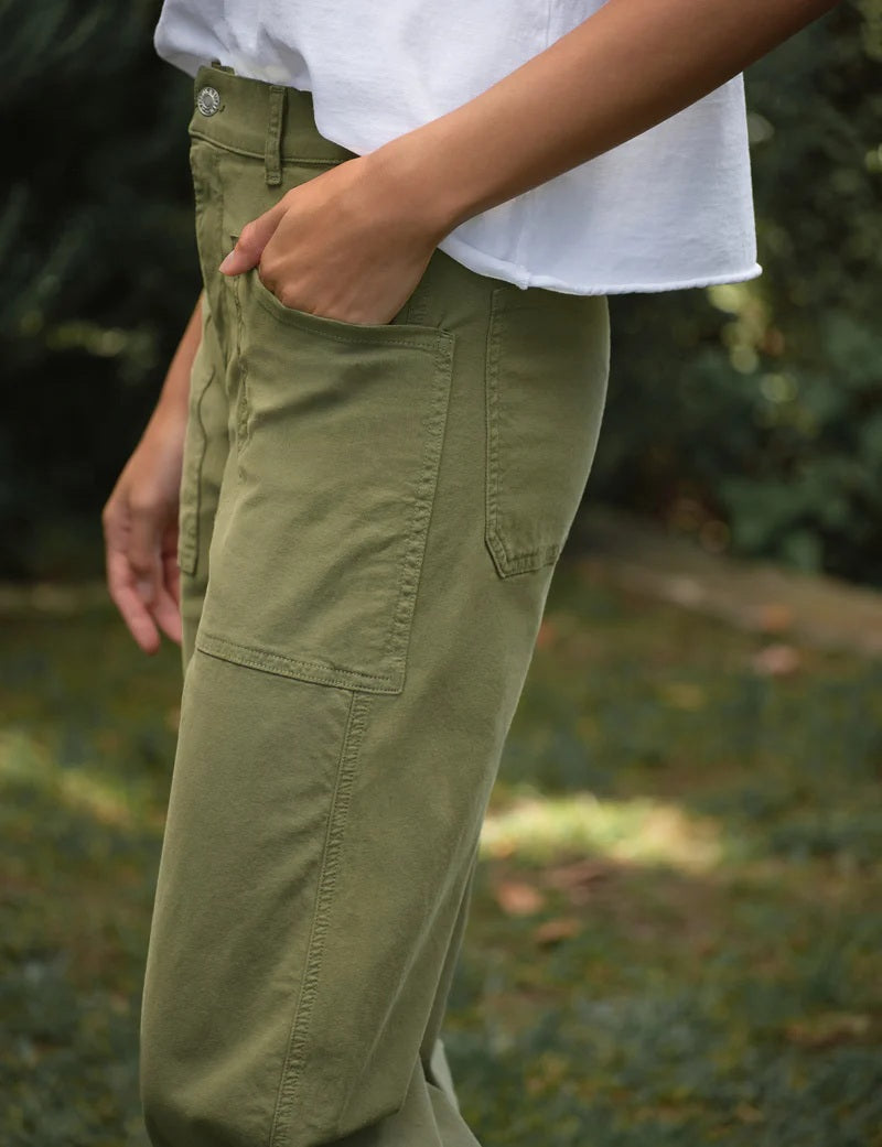Standing on grass surrounded by greenery, a person wears the FRANK &amp; EILEEN BRAY Everyday Barrel Pant in Italian Peached Twill Olive with a cropped fit, paired with a white shirt and beige sandals, hands tucked in their pockets.
