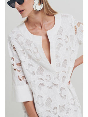 A woman wears the MILVA MI - EMBROIDERED COTTON DRESS IN WHITE by MILVA, styled with bold black and white earrings and black sunglasses. Her straight blonde hair and hand on her hip complete a chic summer resort look.