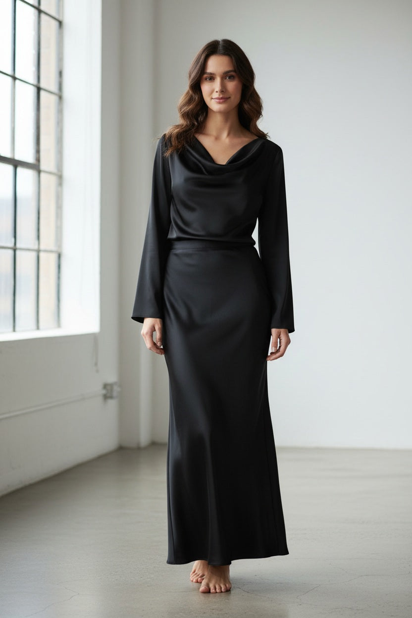 A woman stands against a light gray background, wearing the MILVA MI Satin Blouse in Black by MILVA. The blouse features long sleeves and a draped cowl neckline, and she pairs it effortlessly with wavy brown hair and a slight smile, exuding relaxed sophistication with its elegant silhouette.