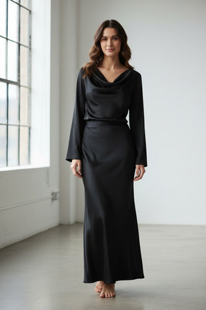 A woman stands against a light gray background, wearing the MILVA MI Satin Blouse in Black by MILVA. The blouse features long sleeves and a draped cowl neckline, and she pairs it effortlessly with wavy brown hair and a slight smile, exuding relaxed sophistication with its elegant silhouette.