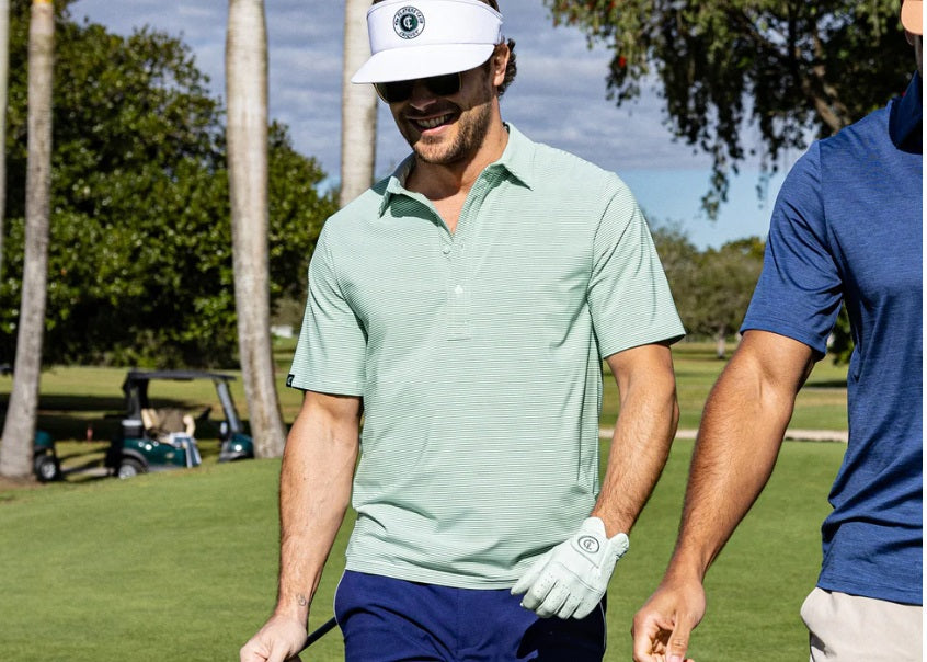 A man wears the CRIQUET Performance Sport Range Polo in Hobby Stripe - Gator, featuring light green and white stripes, a collar, and buttons, standing with one hand in his pocket against a plain background.